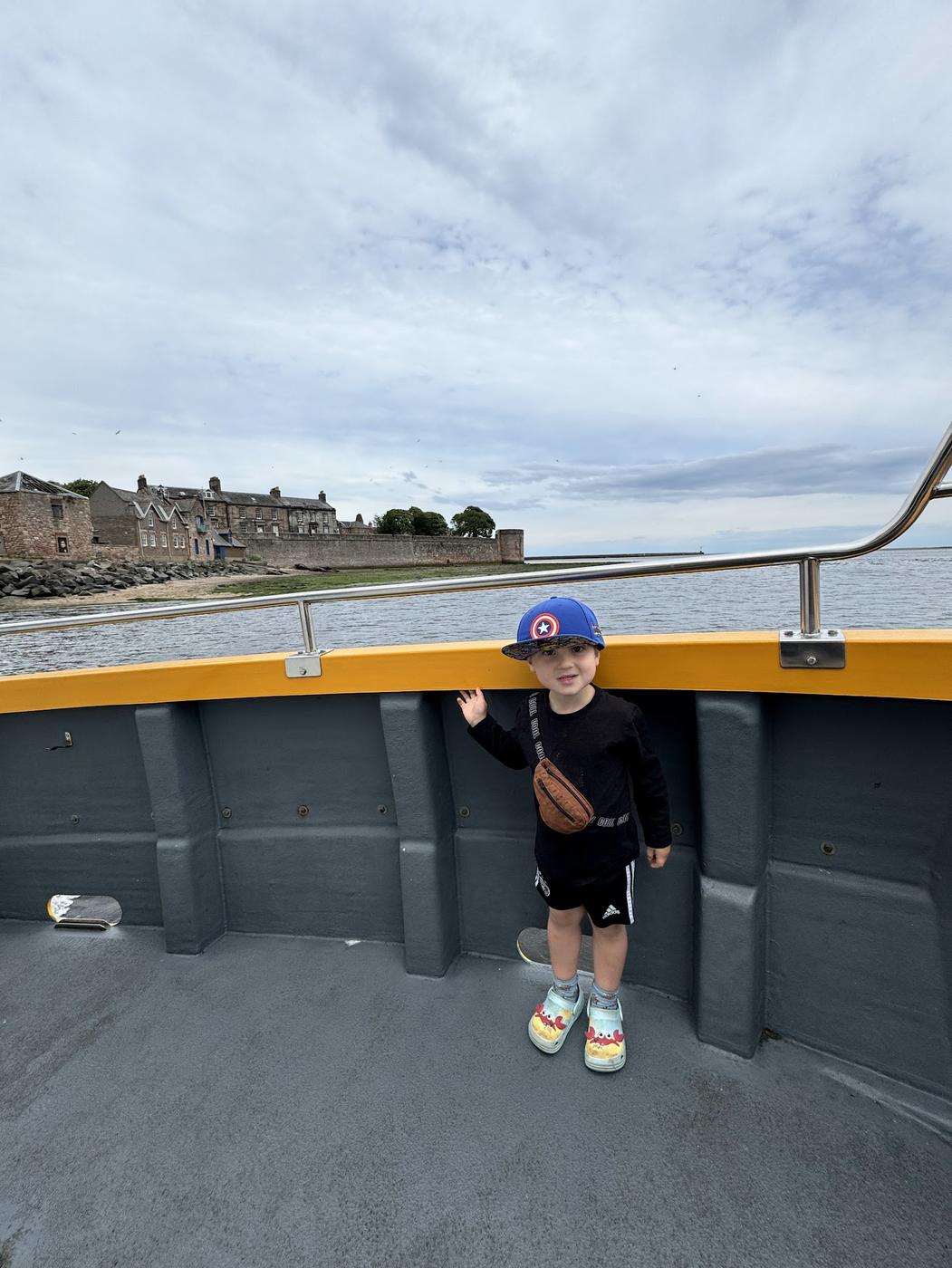A small boy in a cap on the deck of a boat, with Berwick's walls and houses behind.