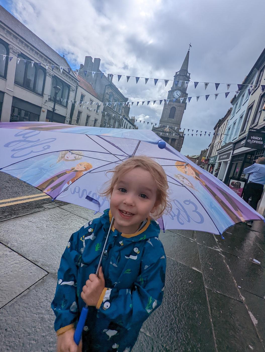 A smiling child with an umbrella on Marygate in Berwick-upon-Tweed, with the Town Hall spire and bunting behind.