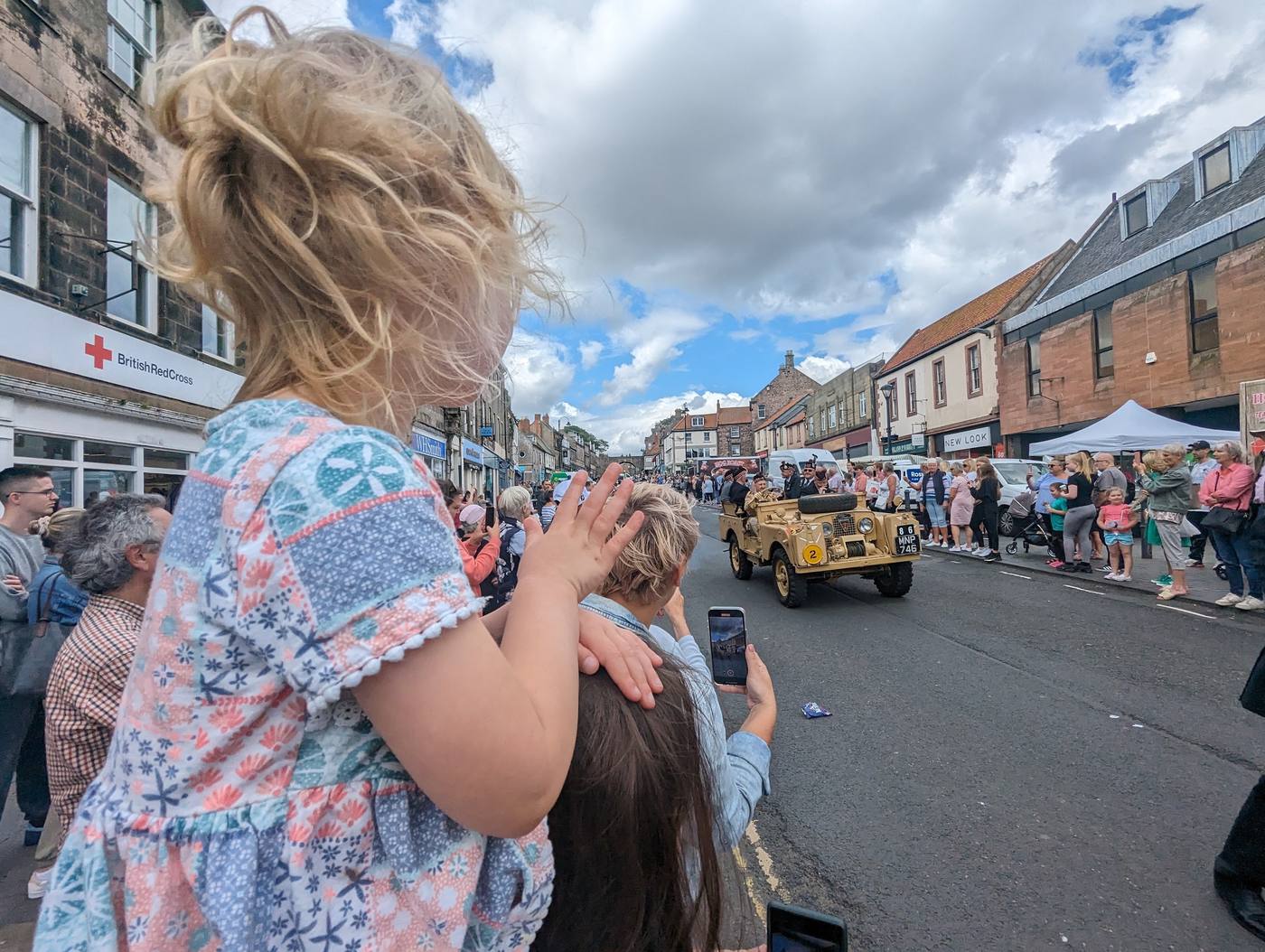 A child with curly hair watching a parade with a vintage Land Rover coming up a crowded street in Berwick.