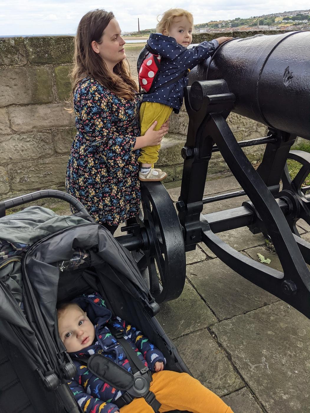 A toddler held up to an old black cannon on Berwick's ramparts, with a baby watching from a buggy.
