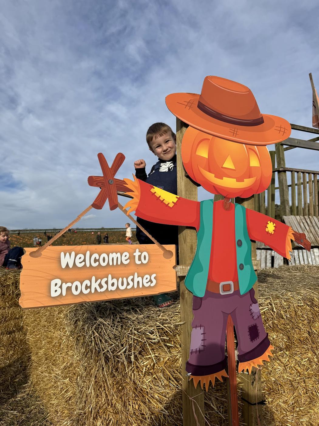 A small boy peeping out behind a smiling pumpkin scarecrow that says Welcome to Brocksbushes, on a pile of straw bales.