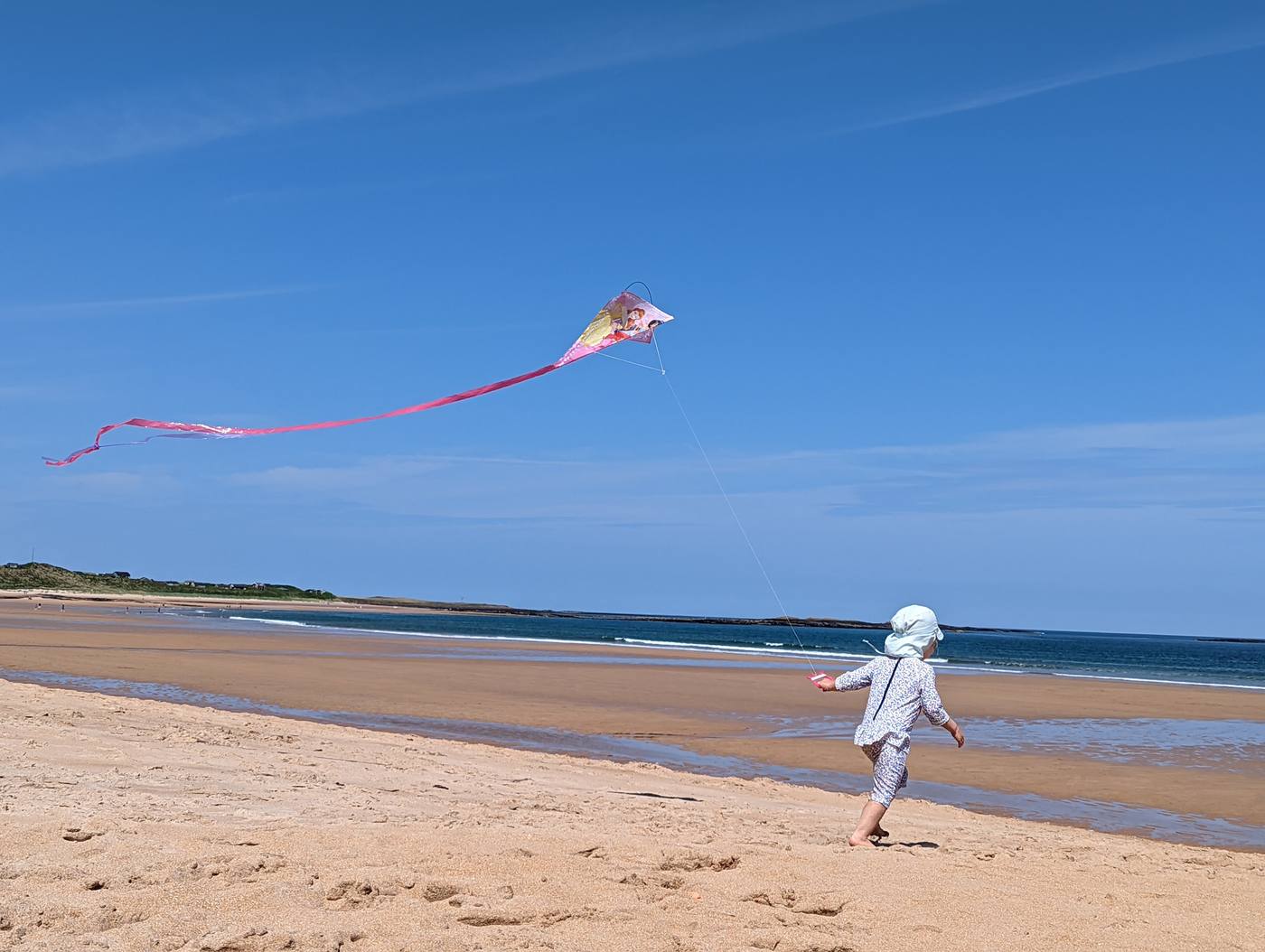 A small child in a sun hat running with a pink kite across the wide sands of Embleton Bay.