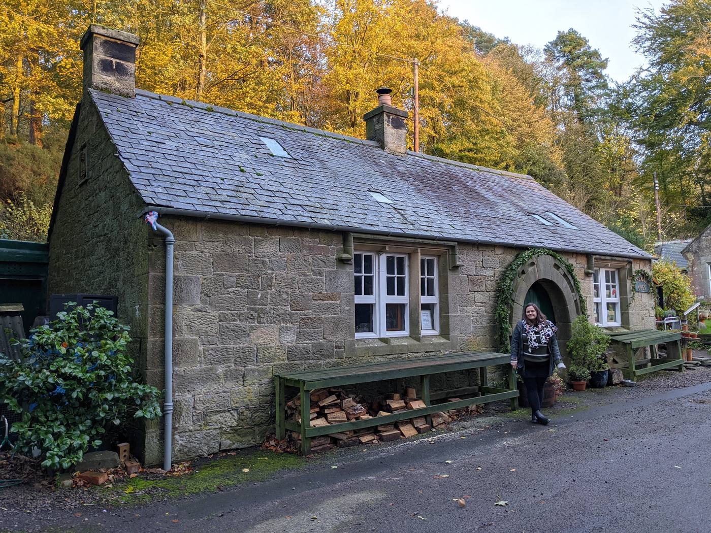 A stone cottage with white windows and a wood store, under golden autumn trees in Ford village.