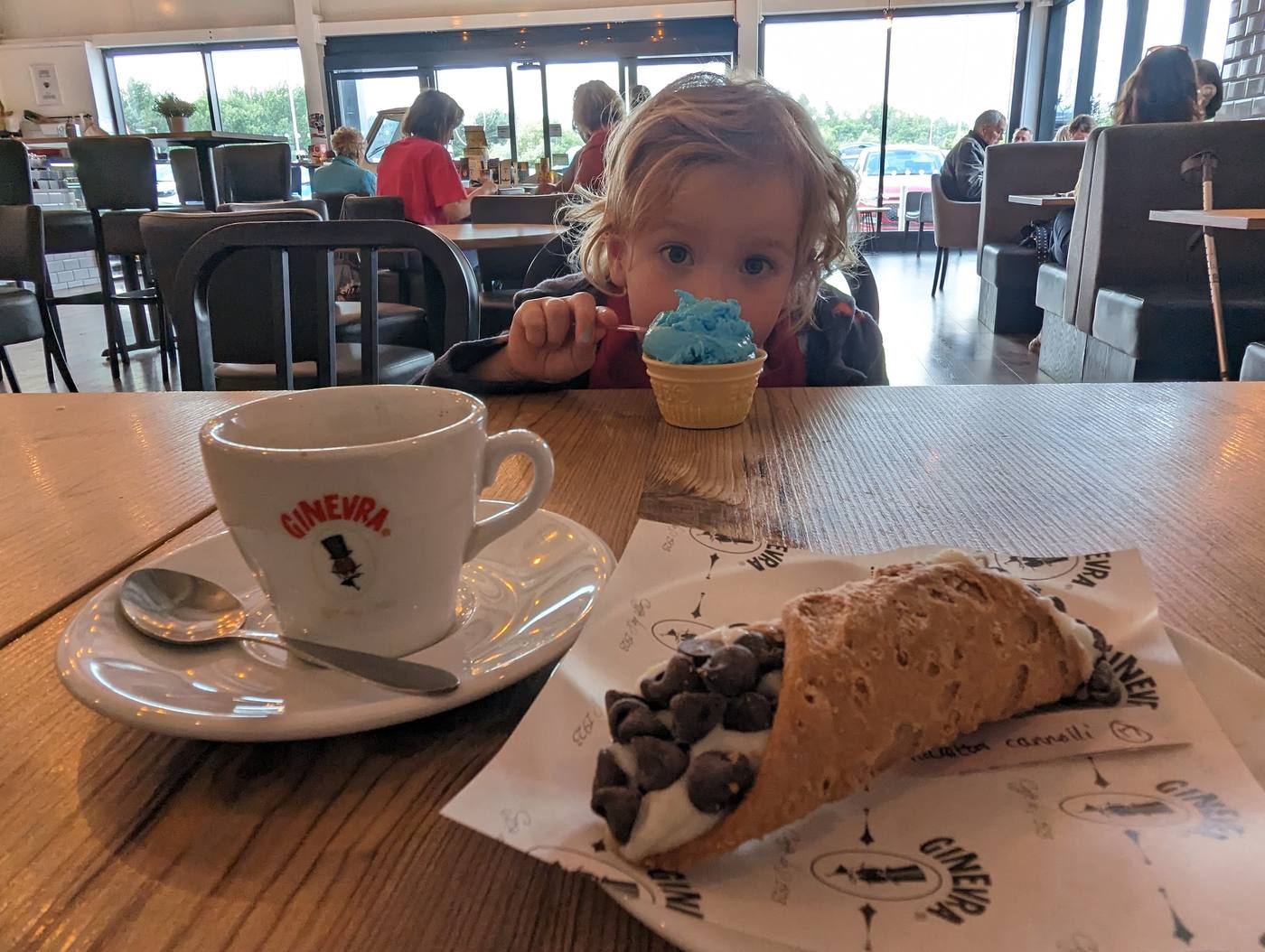 A coffee and a chocolate chip cannolo on the table, with a child eating a blue ice cream behind, at Ginevra in Prudhoe.