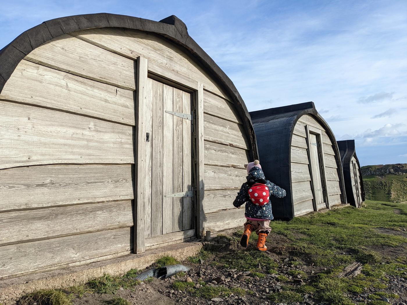 A toddler in a red backpack walks past the black-roofed upturned-boat sheds on Holy Island.
