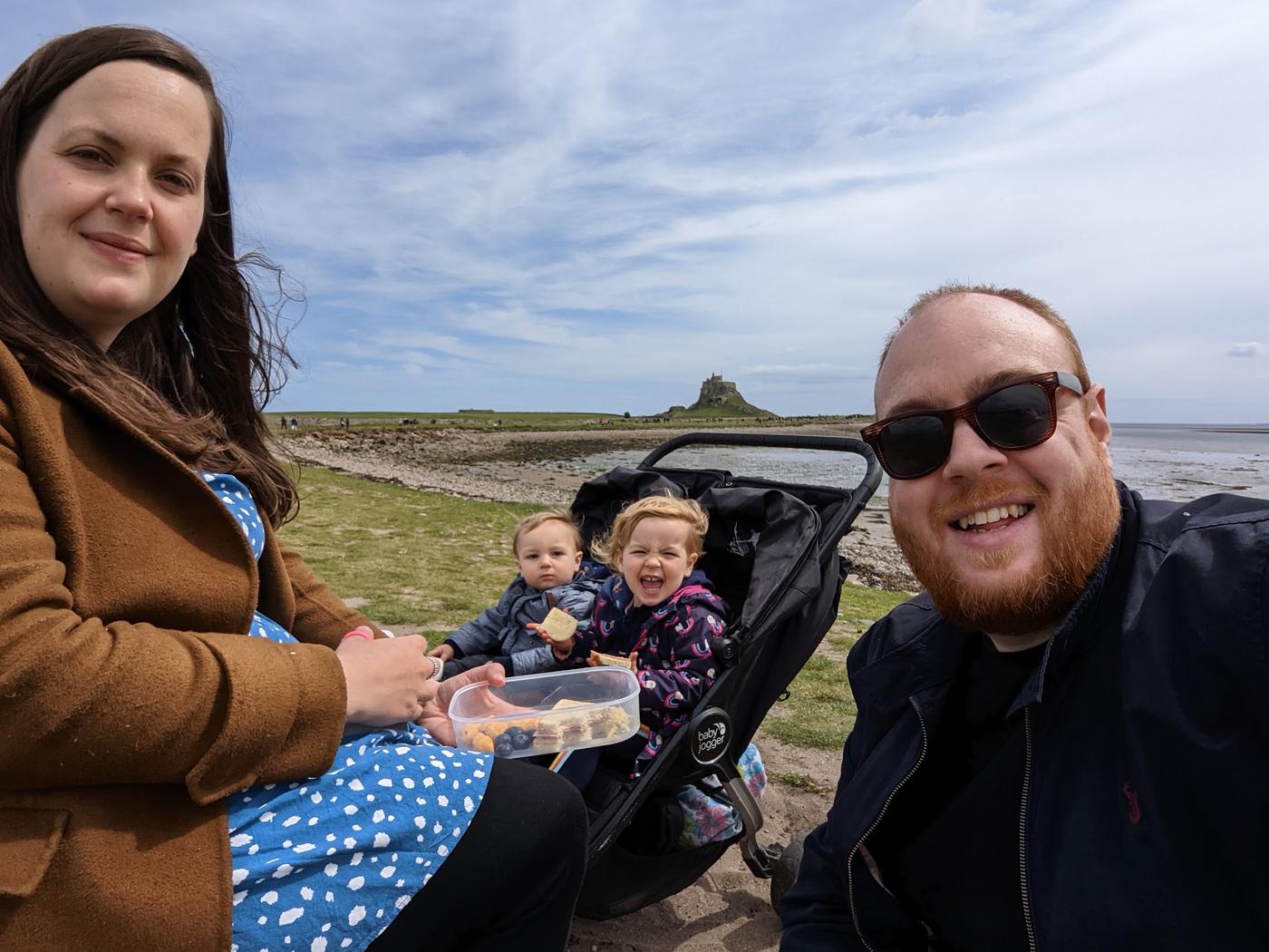 Dan, Julia and their two small children picnicking on the grass on Holy Island, with Lindisfarne Castle in the distance.
