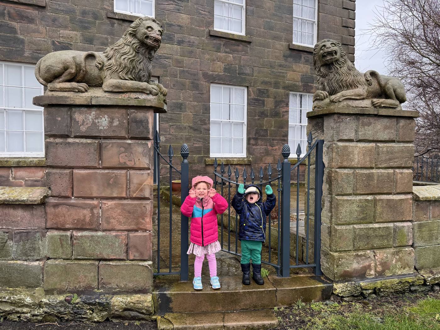 Two children in winter coats cheering at the iron gate of Lion House in Berwick, with a stone lion on each gatepost.