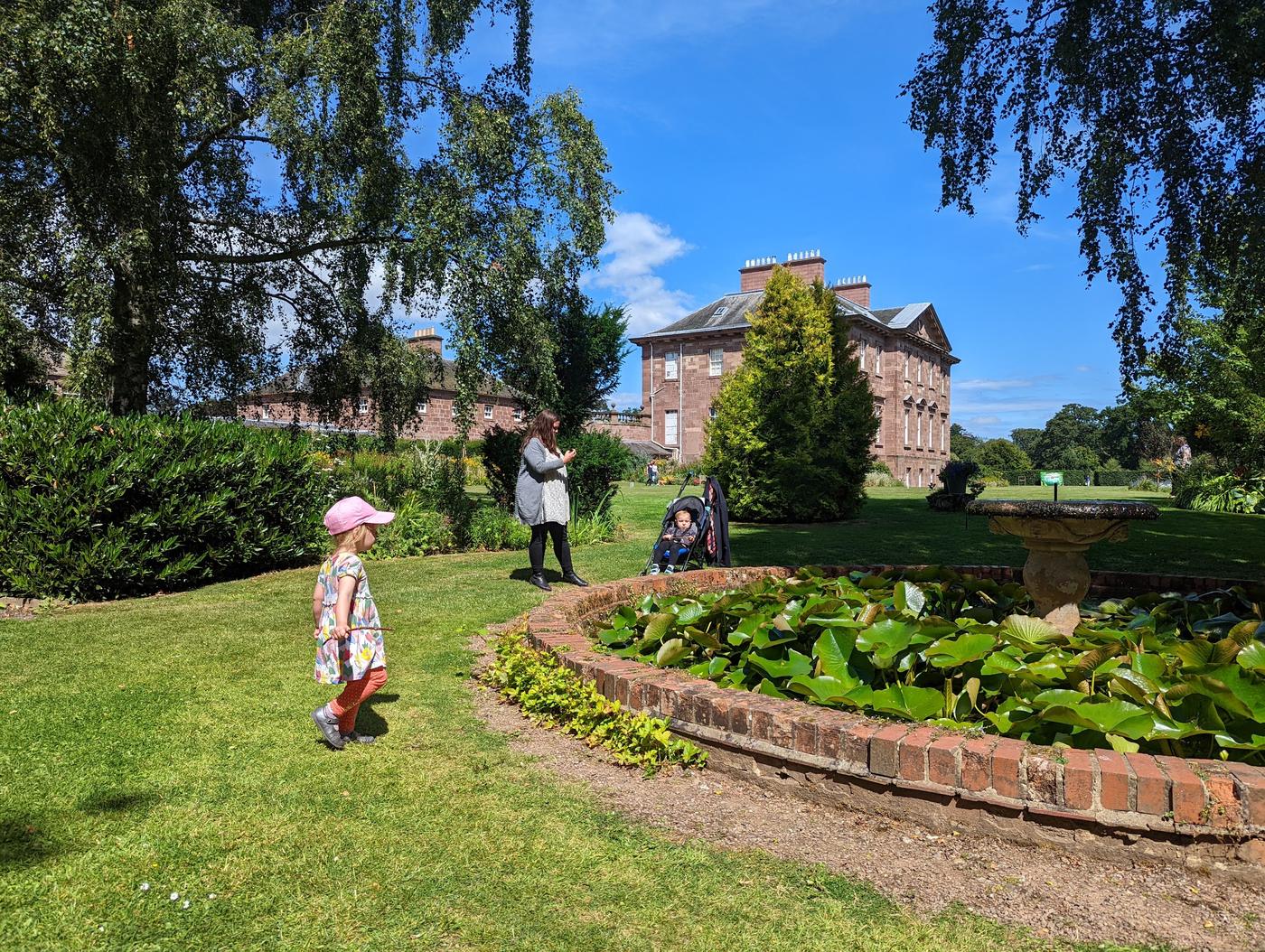 A child in a sun hat on the lawn by the lily pond at Paxton House, with the Georgian house behind.
