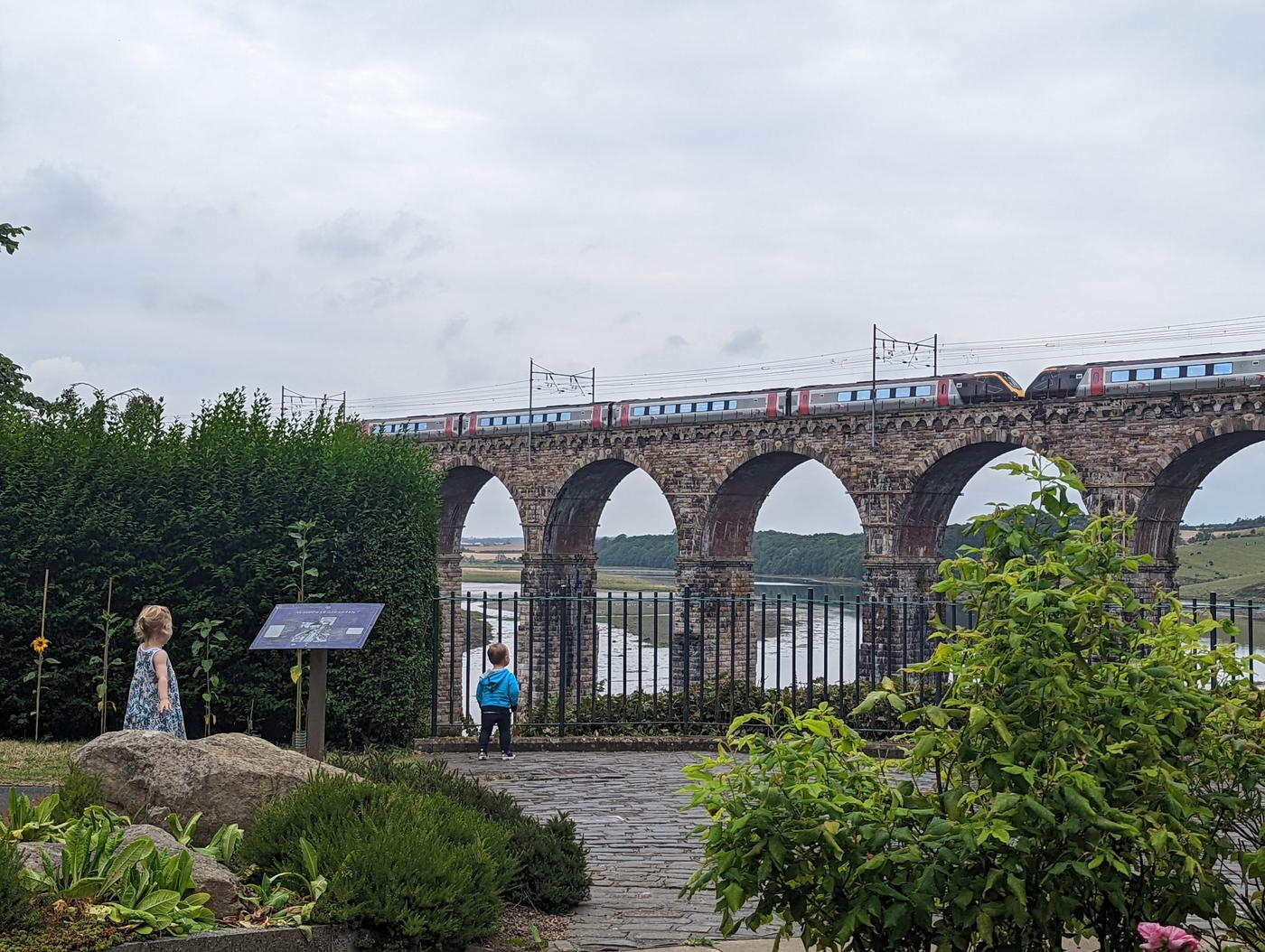 Two children in a riverside garden watching a train cross the arches of the Royal Border Bridge.