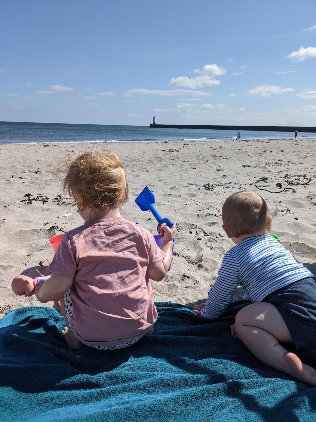 Two small children on a towel on Spittal beach, with Berwick's pier and lighthouse on the horizon.