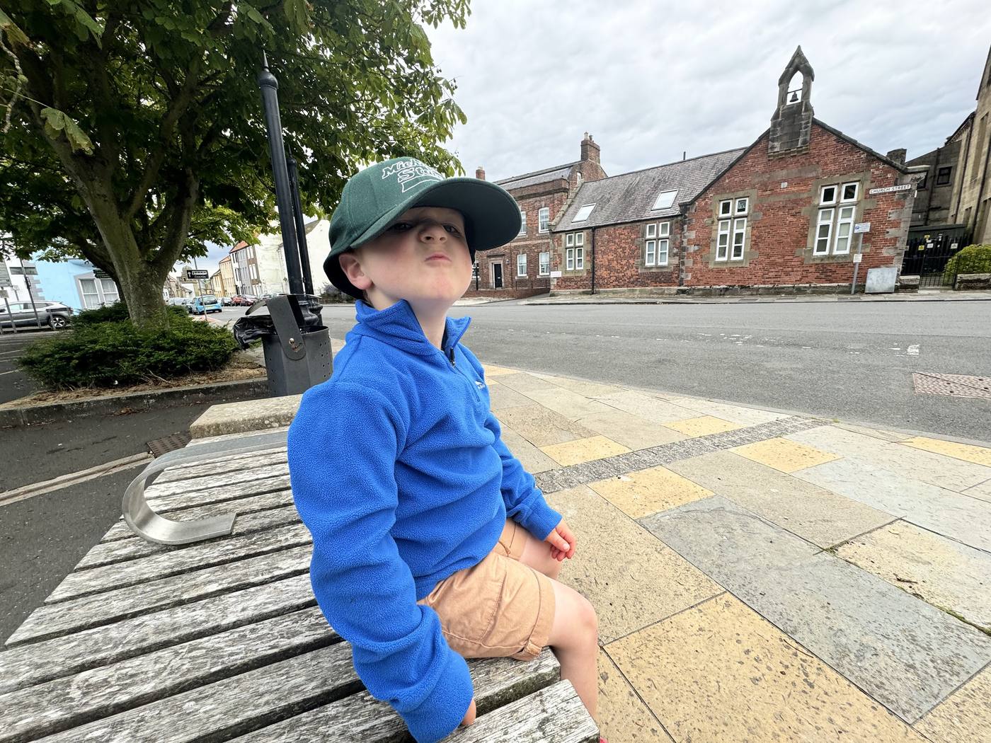 A boy in a green cap and blue fleece sitting on a bench on the Parade in Berwick, by an old red-brick building with a bell.