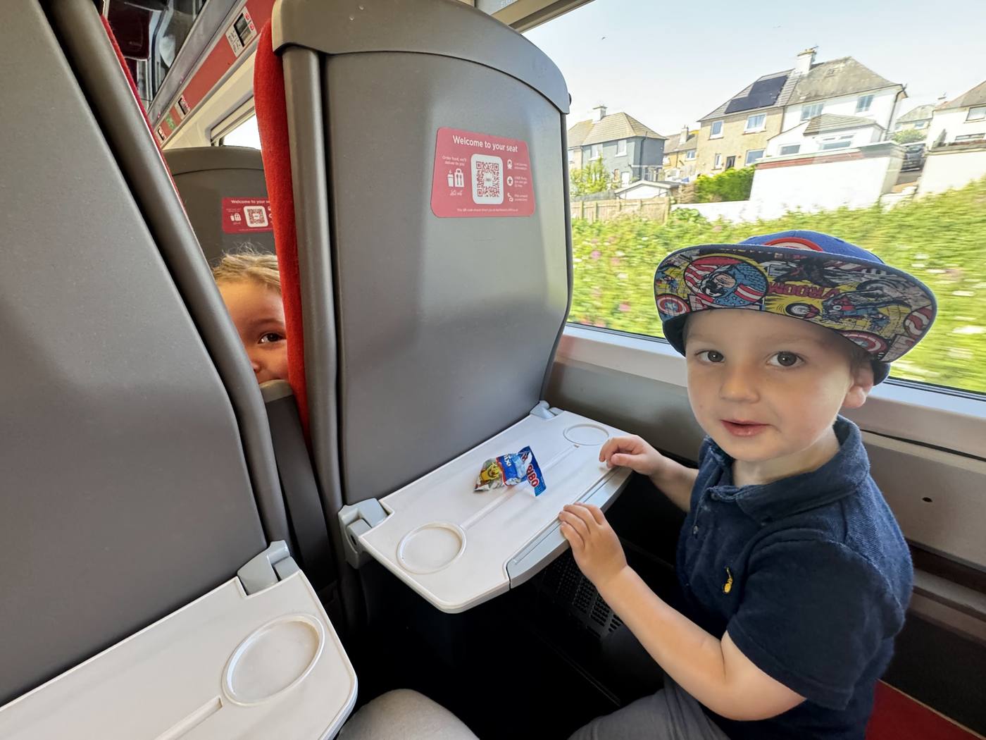 A boy in a colourful sun hat at a train window seat, with his sister peeping round the seat behind.