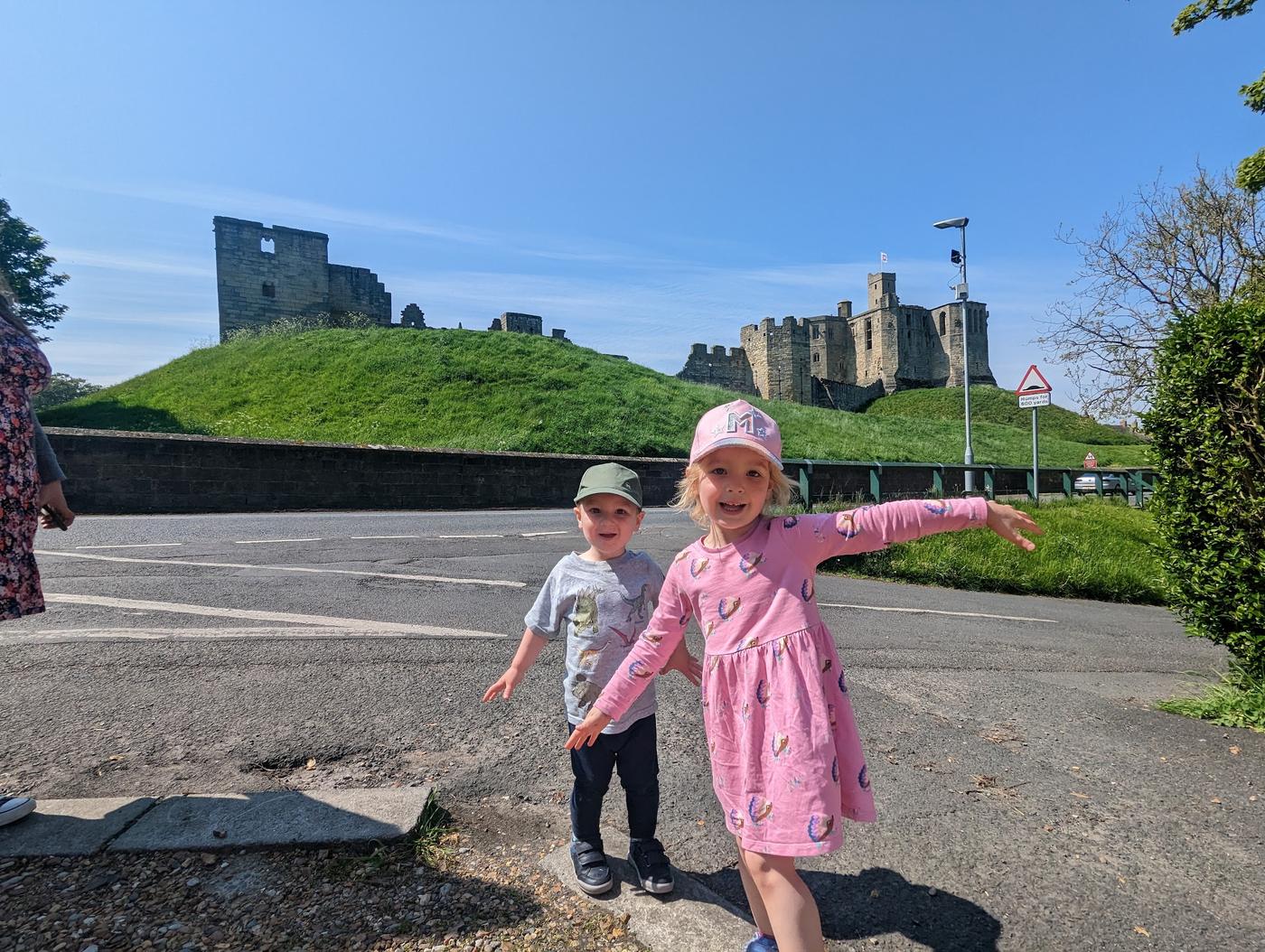 Two children posing on the pavement below Warkworth Castle on its grassy mound.