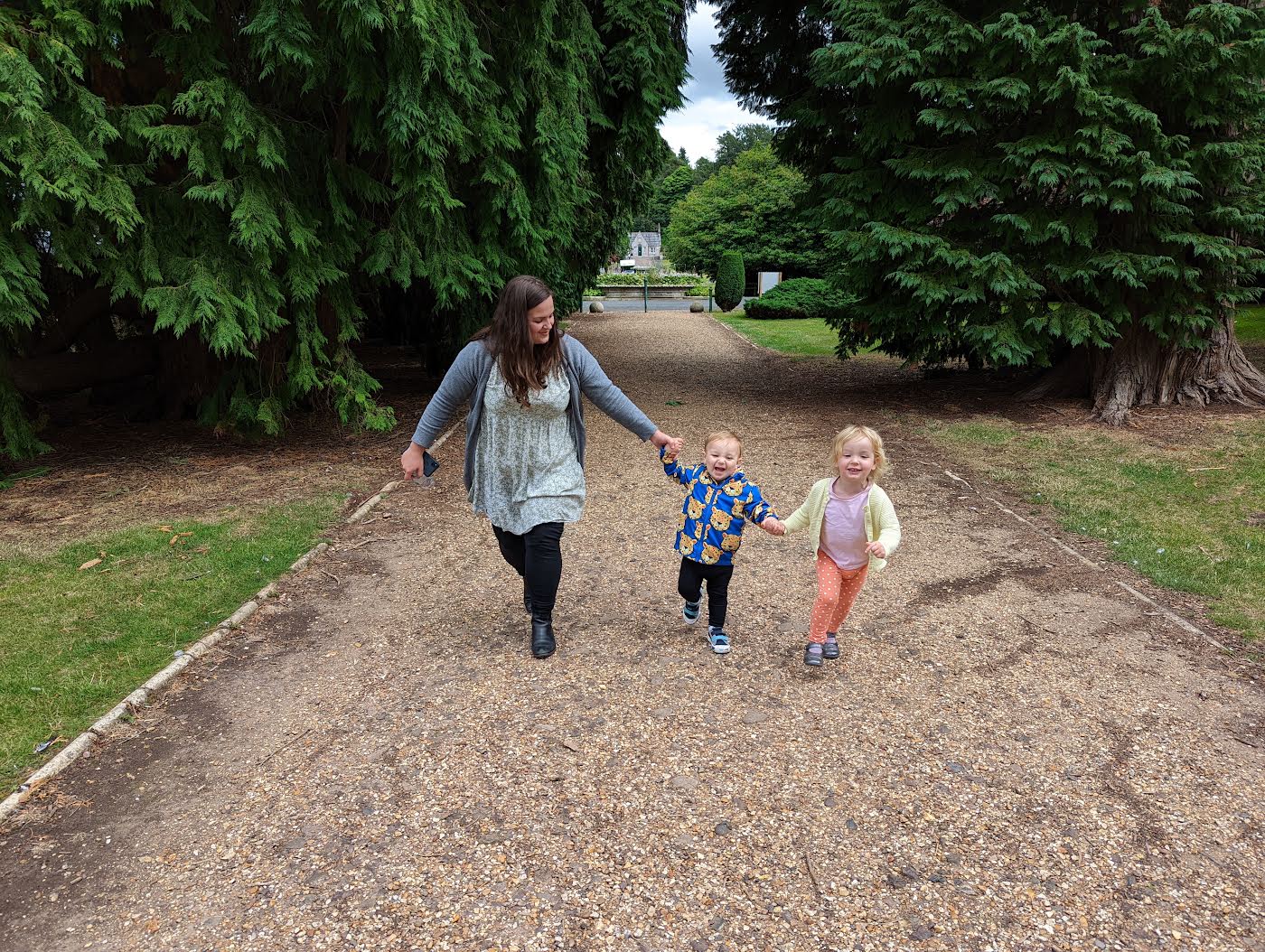 A woman walking hand in hand with two small children down a gravel lane between tall trees and lawns in Ford village.